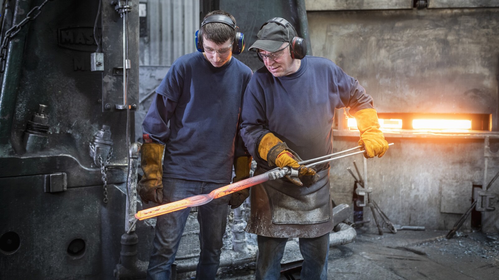 En äldre man vägleder en yngre i stålindustri