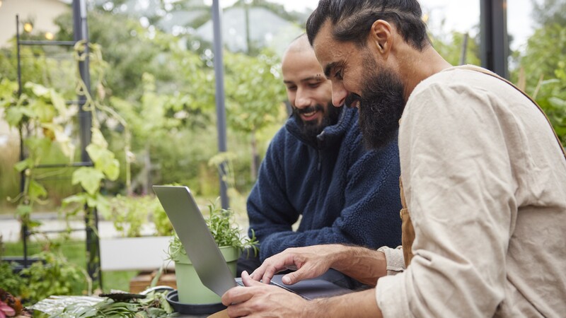 Two men sit in a greenhouse with a laptop